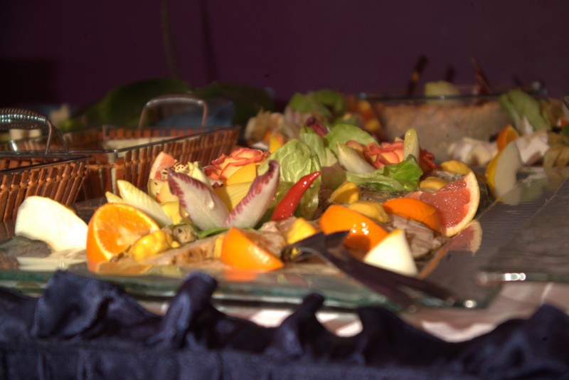 Szklany półmisek z dekoracyjnie ułożonymi kawałkami owoców i warzyw, w tym pomarańcze, grejpfruty, sałata i papryczka chili, na tle bufetu z koszykami i innymi naczyniami.